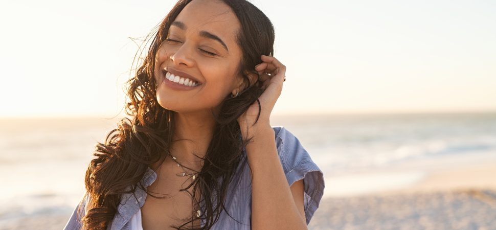 Smiling latin hispanic woman relaxing on beach with closed eyes at sunset. Beautiful mixed race woman enjoying wind fluttering hair. Charming and calm young woman breathing fresh air at summer beach with copy space.